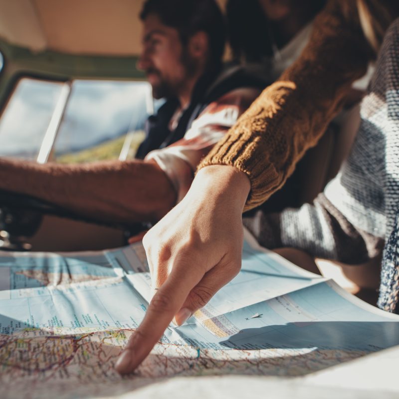 Friends on road trip looking at map for directions. Close up of female pointing at the map while man driving van in background.